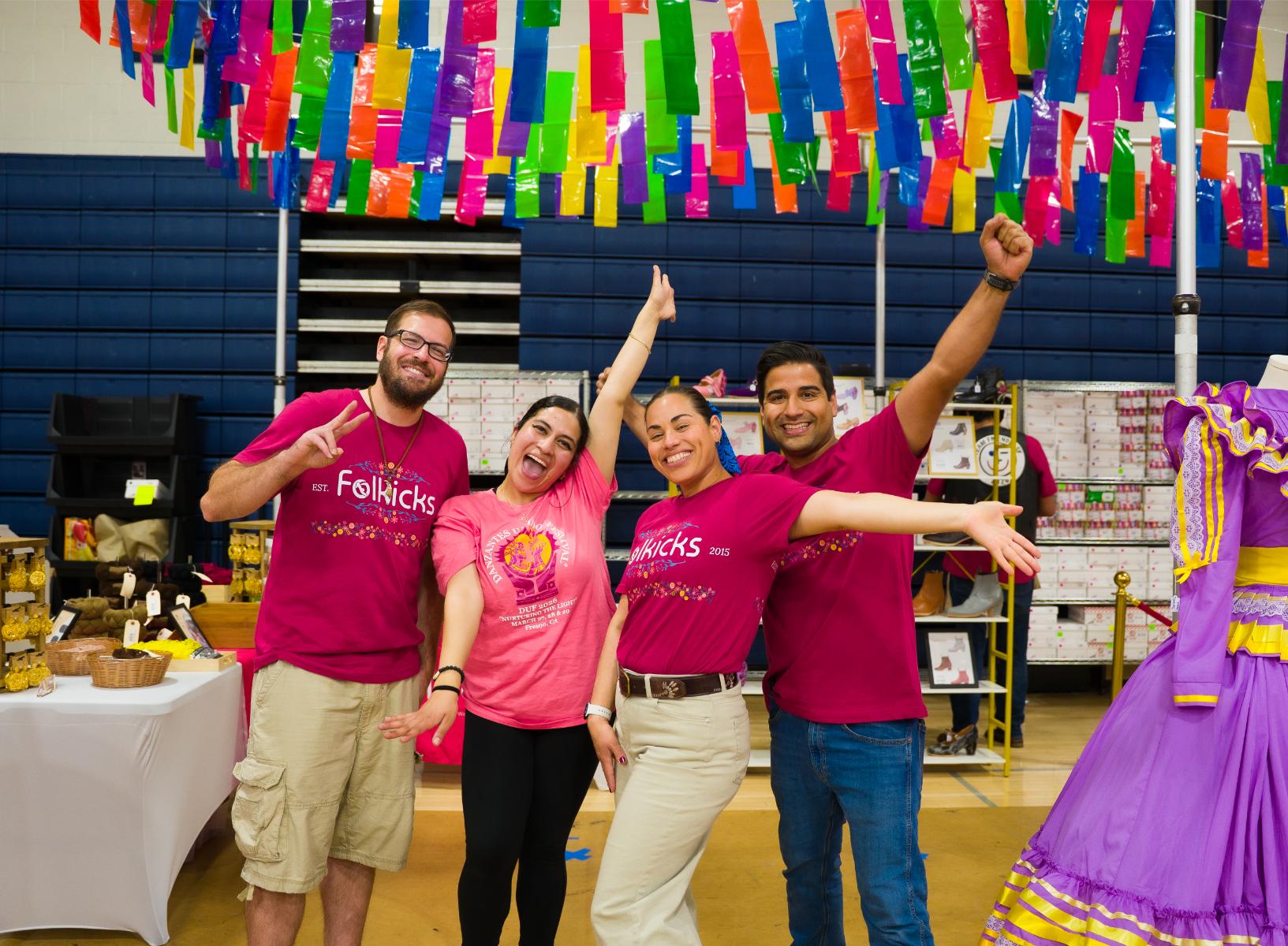 The Folkicks team smiling and celebrating at a dance festival booth, wearing branded Folkicks t-shirts, with shoe displays and colorful decorations in the background.