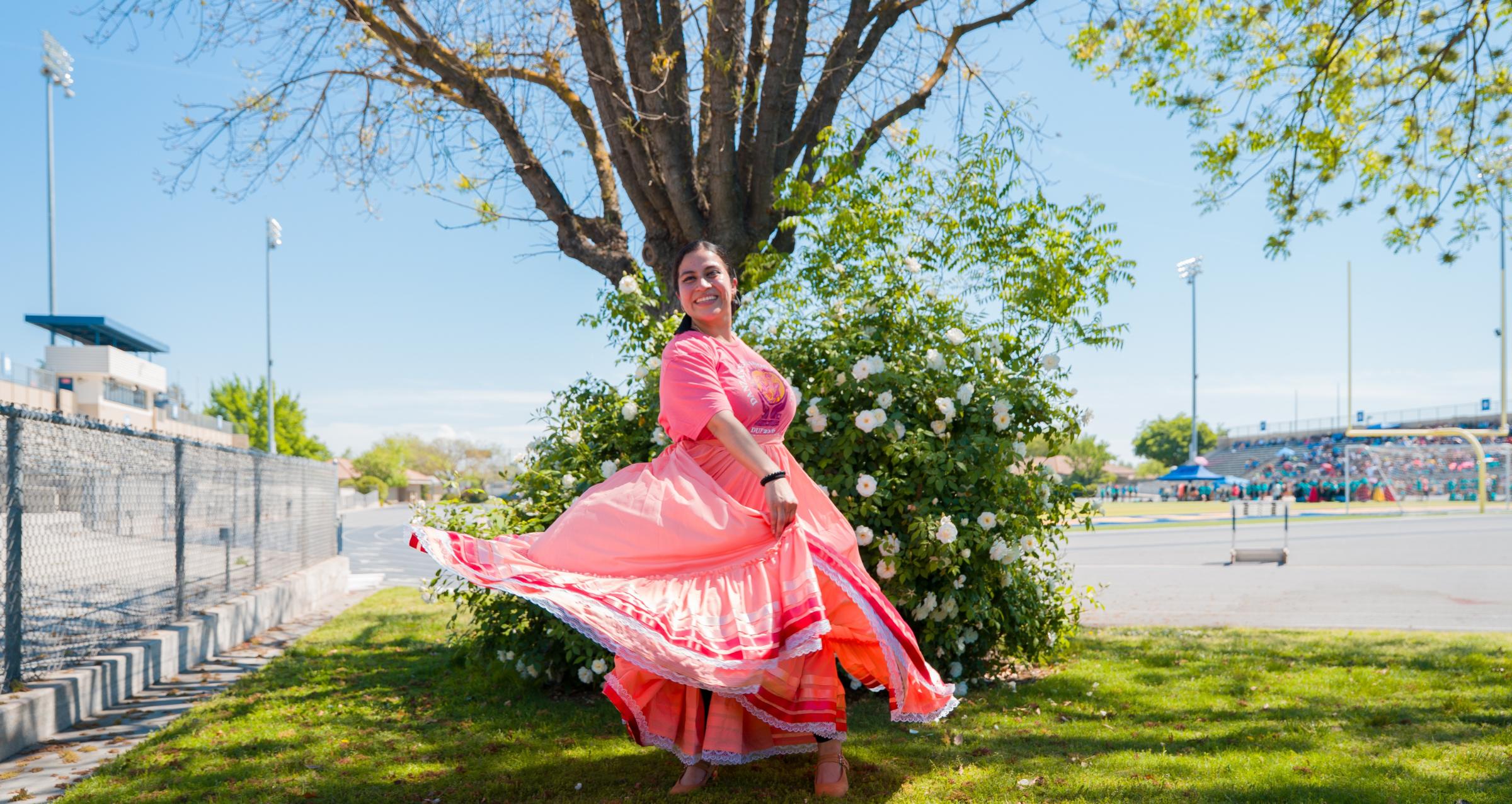 A dancer twirling in a pink and coral folklórico skirt with striped and lace trim, smiling in front of a blooming white rose bush.