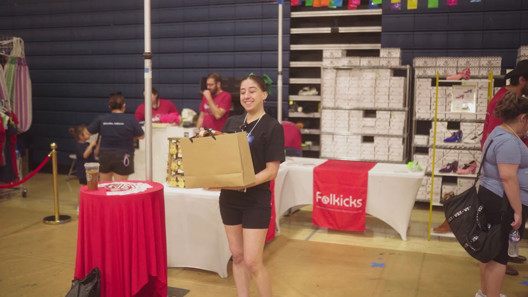 Video of a Folkicks staff member at a warehouse event, smiling while holding up a shopping bag, with shelves of shoeboxes and a Folkicks banner in the background.