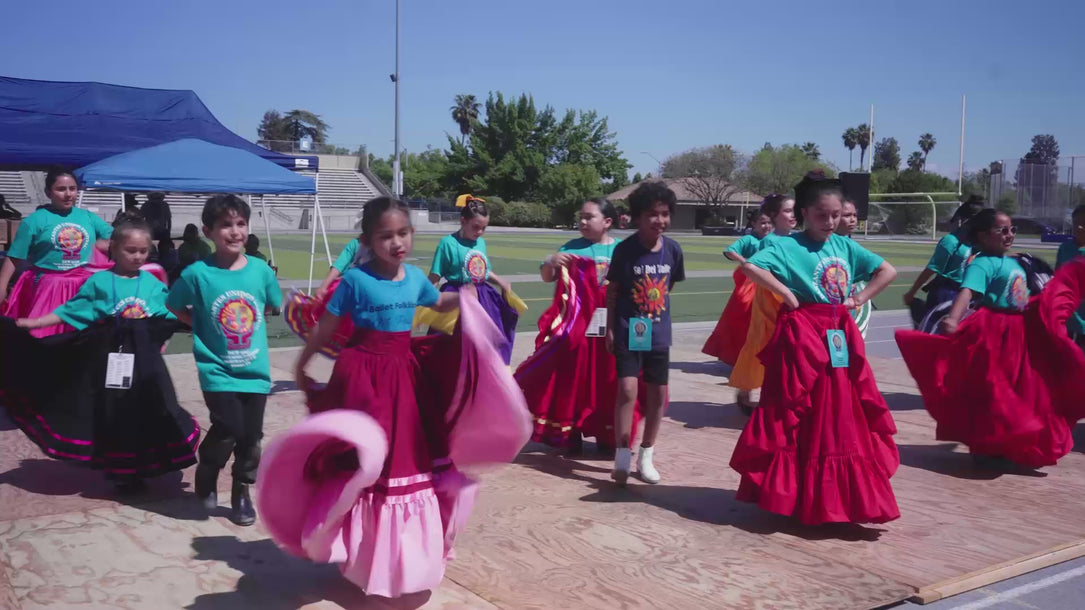 Video of school-age folklórico dancers performing outdoors on a wooden stage, wearing team t-shirts and swirling vibrant skirts in pink, red, and purple.